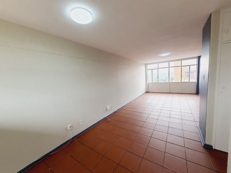 A spacious, empty living area featuring terracotta floor tiles, large windows providing natural light, and neutral wall finishes.