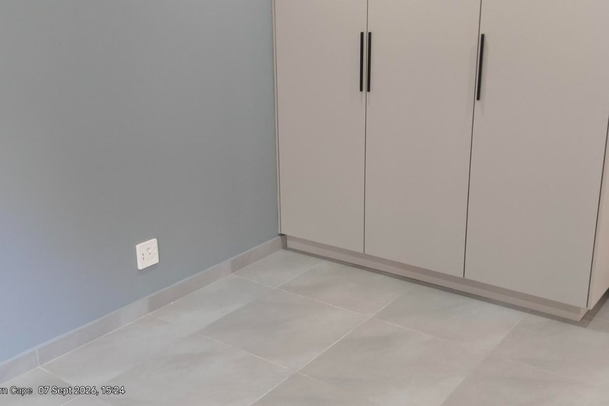A clean, modern bedroom featuring neutral grey walls, large format floor tiles, and a built-in wardrobe with contemporary black handles.