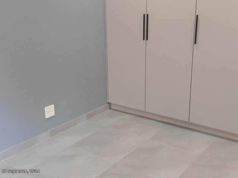 A clean, modern bedroom featuring neutral grey walls, large format floor tiles, and a built-in wardrobe with contemporary black handles.