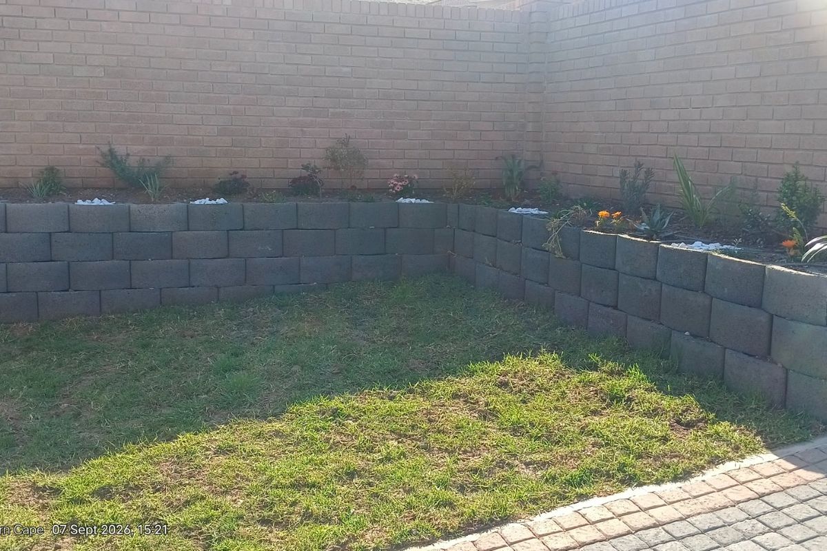 A private garden area featuring a brick boundary wall, tiered retaining wall planters, and a small lawn section adjacent to a paved driveway.