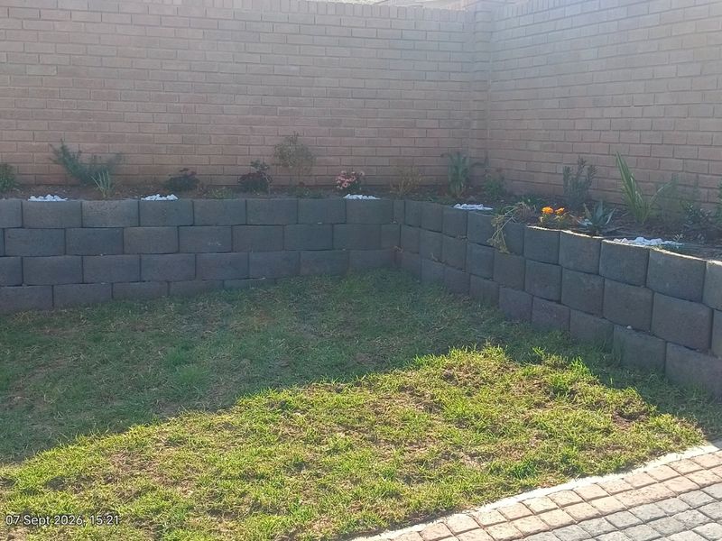 A private garden area featuring a brick boundary wall, tiered retaining wall planters, and a small lawn section adjacent to a paved driveway.