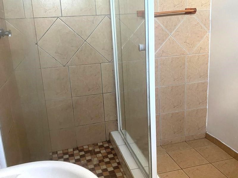 A functional bathroom featuring a glass shower enclosure with tiled flooring, a pedestal basin, and neutral wall tiling.