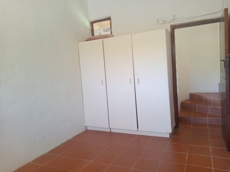 A simple bedroom featuring tiled flooring, a built-in wardrobe, and a small high-level window, leading to an adjacent staircase.