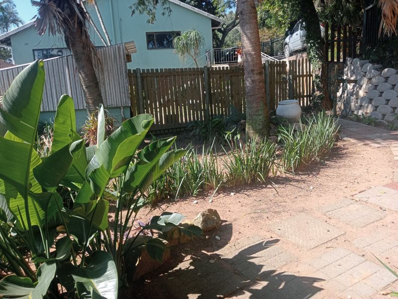 A garden area featuring a wooden fence, a stone retaining wall, and a paved walkway surrounded by established plants and trees.