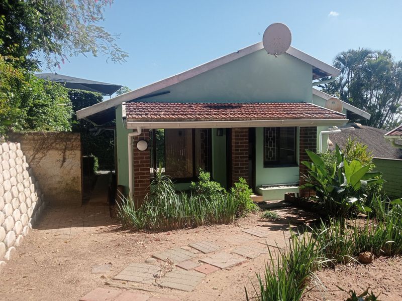 Front elevation of a single-story apartment featuring a terracotta tiled roof, brick pillar accents, and a paved entrance path with surrounding garden.