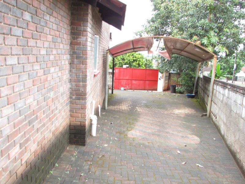 Rear exterior area featuring a paved driveway, brick walling, and a damaged carport structure leading to a gated entrance.