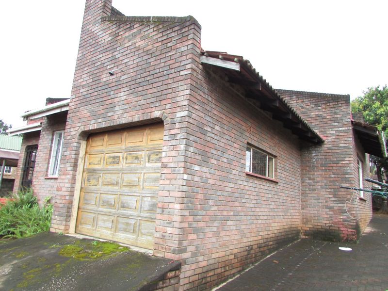 Exterior view of a brick house featuring a garage with a weathered door and a paved driveway with visible moss growth.