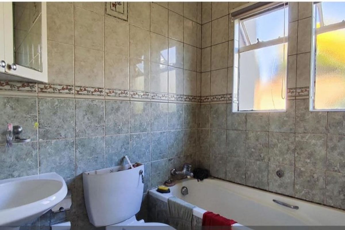 A functional bathroom featuring a bathtub, pedestal basin, and toilet, finished with dated wall tiling and natural light from dual casement windows.