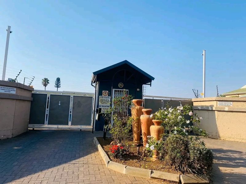 Secure apartment complex entrance featuring a gated access point, guard house, paved driveway, and perimeter wall with electric fencing.