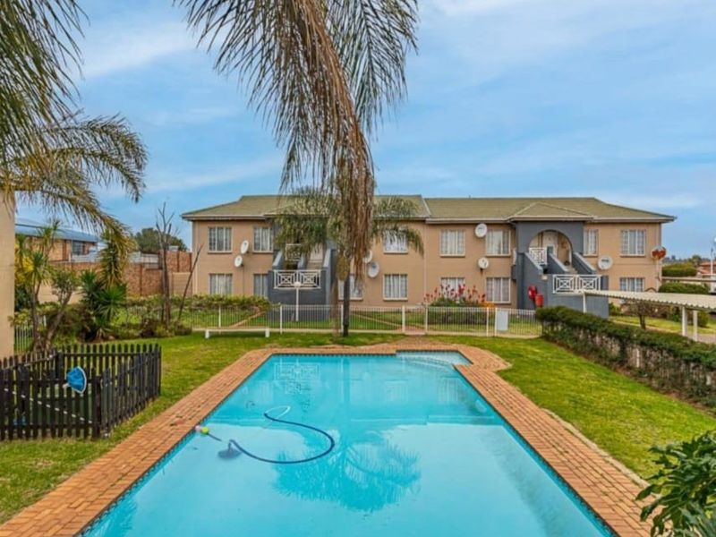 A well-maintained communal swimming pool area set within a landscaped garden, featuring brick coping and views of the apartment complex facade.