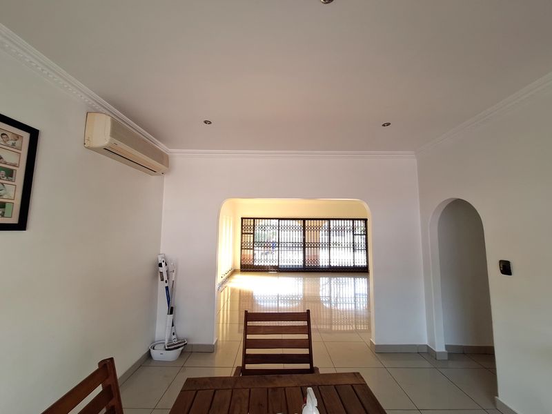 A bright living area featuring tiled flooring, recessed downlights, decorative ceiling cornices, and a split-system air conditioner, leading through an archway to a secure patio.