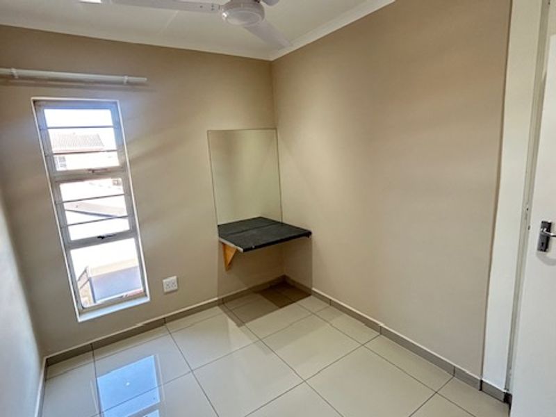 A clean, empty bedroom featuring tiled flooring, a ceiling fan, and a built-in desk with a wall-mounted mirror.