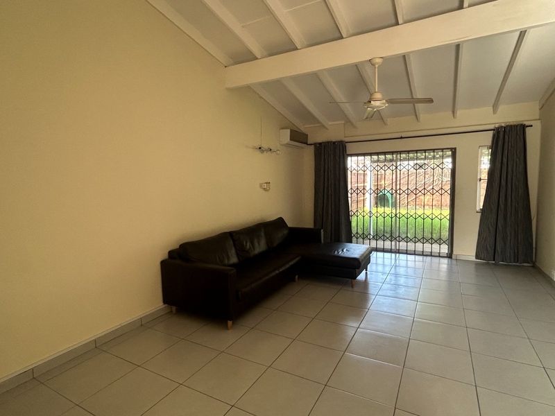 Spacious living room featuring tiled flooring, exposed beam ceiling, ceiling fan, air conditioning, and direct access to the rear garden through a security gate.