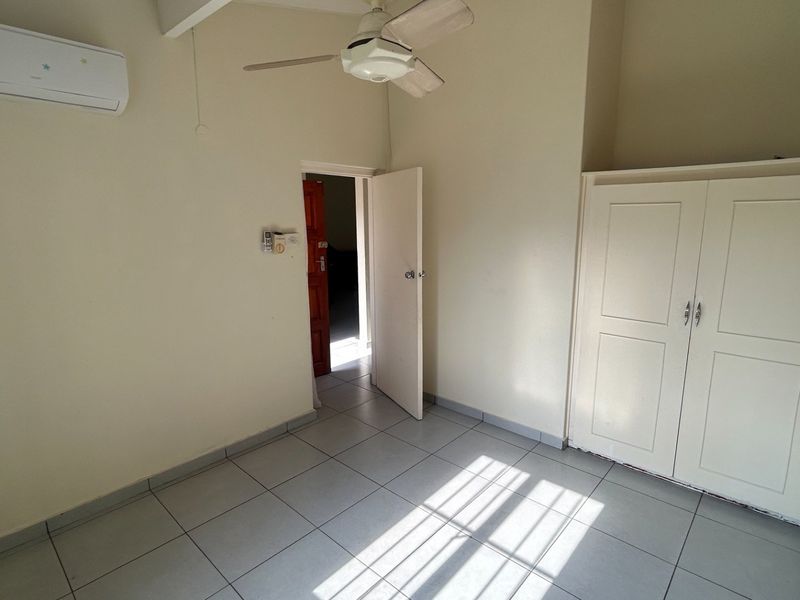 A bright, empty bedroom featuring tiled flooring, a built-in wardrobe, a ceiling fan, and a wall-mounted air conditioning unit.