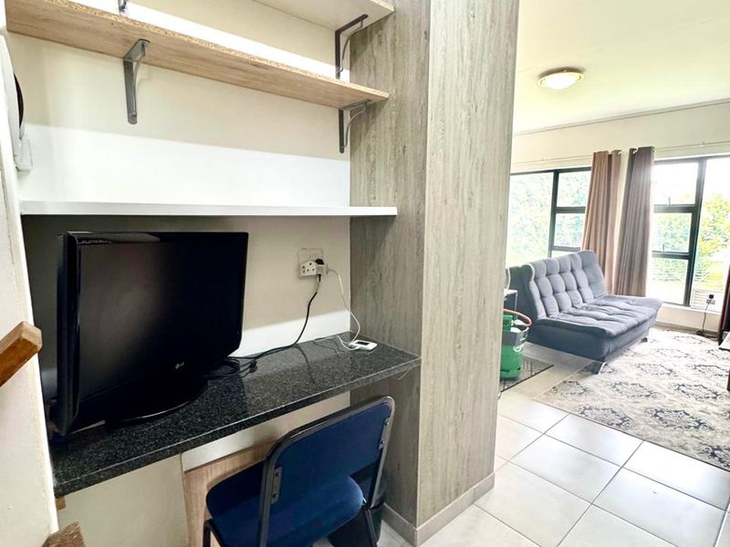 Open plan living area featuring a built-in desk with floating shelving, tiled flooring, and large windows providing natural light.