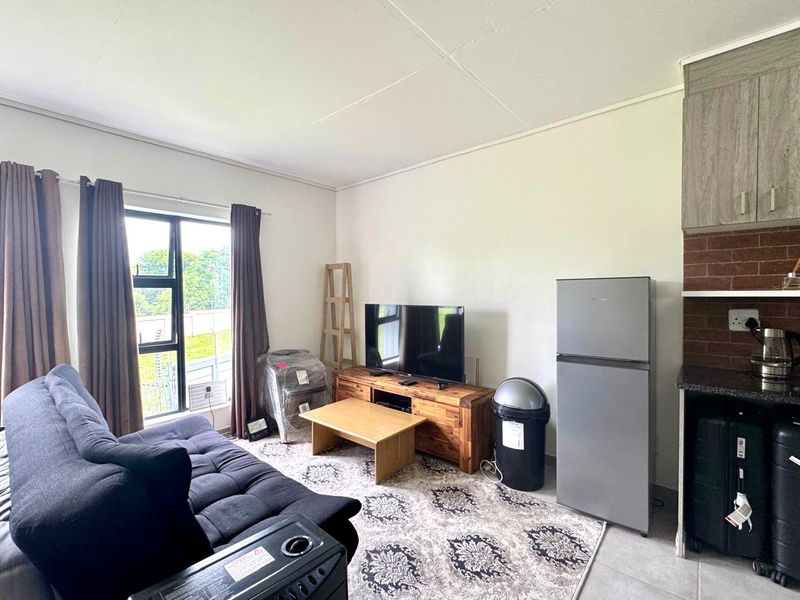 Open-plan living area featuring tiled flooring, aluminium window frames, and a compact kitchen section with exposed brick backsplash and built-in cabinetry.