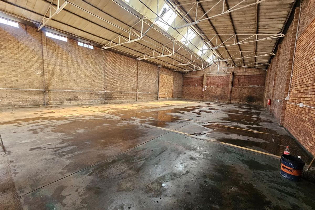 Large industrial warehouse space featuring exposed brick walls, steel roof trusses, and natural light from overhead skylights.