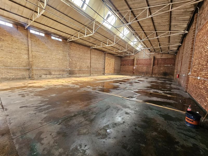 Large industrial warehouse space featuring exposed brick walls, steel roof trusses, and natural light from overhead skylights.