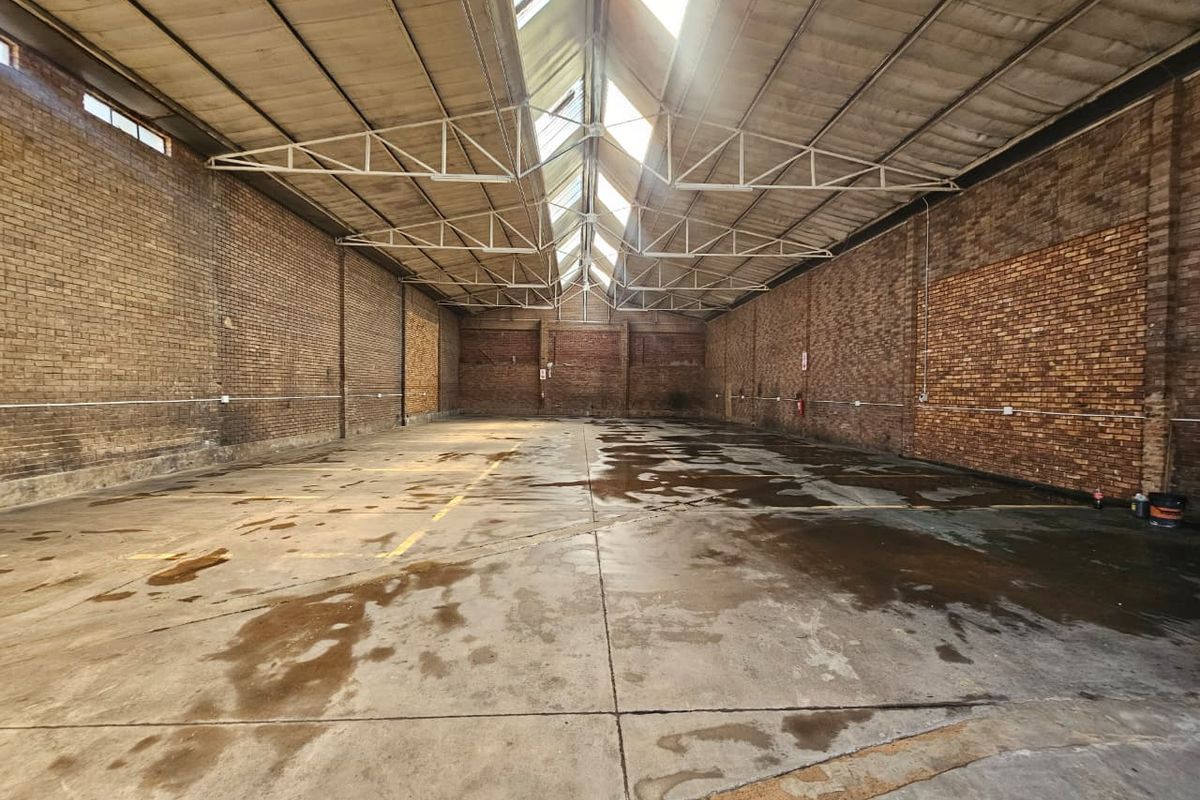 Spacious industrial warehouse unit featuring exposed brick walls, concrete flooring, and a high truss roof with central skylights providing natural light.