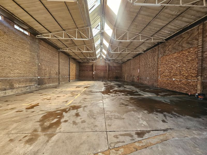 Spacious industrial warehouse unit featuring exposed brick walls, concrete flooring, and a high truss roof with central skylights providing natural light.