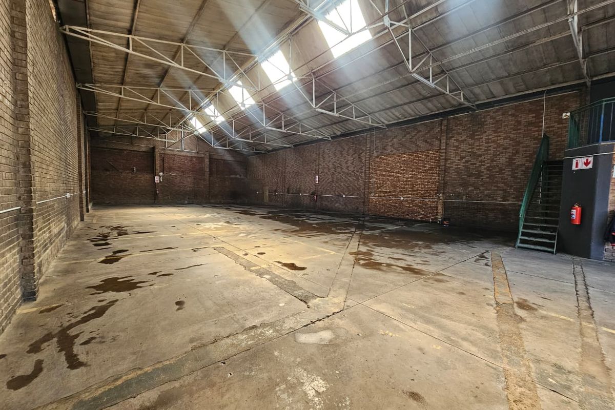 Spacious industrial warehouse unit featuring exposed brick walls, high steel truss ceilings with skylights, concrete flooring, and an integrated metal staircase.