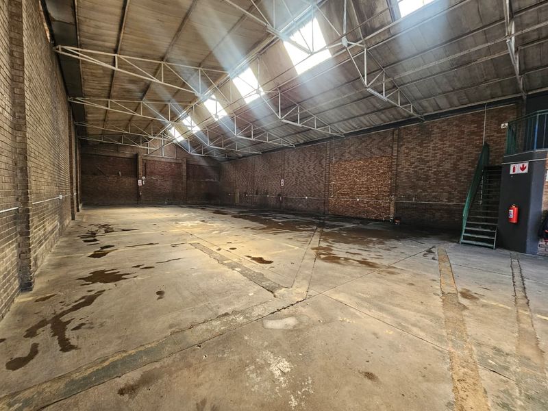Spacious industrial warehouse unit featuring exposed brick walls, high steel truss ceilings with skylights, concrete flooring, and an integrated metal staircase.