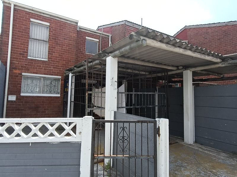 Front exterior view of a brick townhouse featuring a covered carport, security gate, and perimeter walling in need of maintenance.
