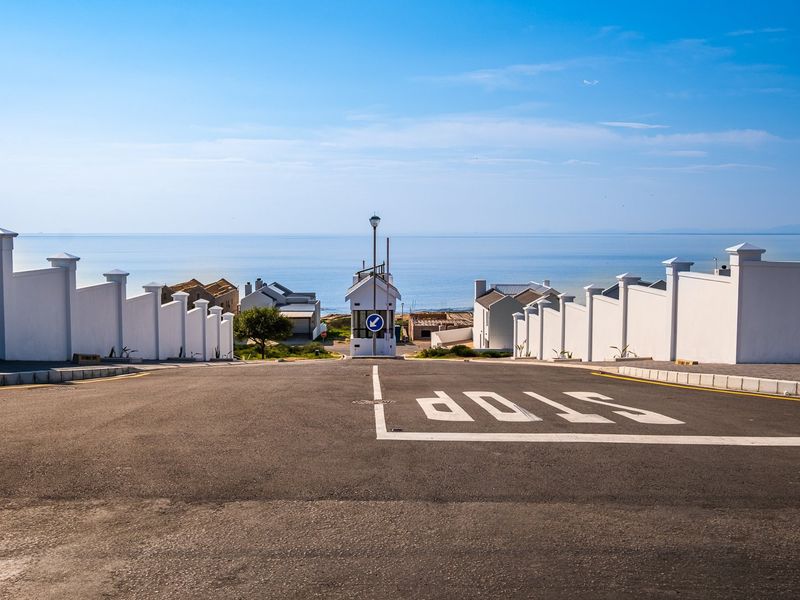 A clear street view looking down a paved road towards a security gatehouse with ocean views in the background.