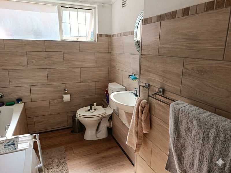 A functional bathroom featuring wood-look wall and floor tiles, a bathtub, a pedestal basin, and a toilet with security bars on the window.