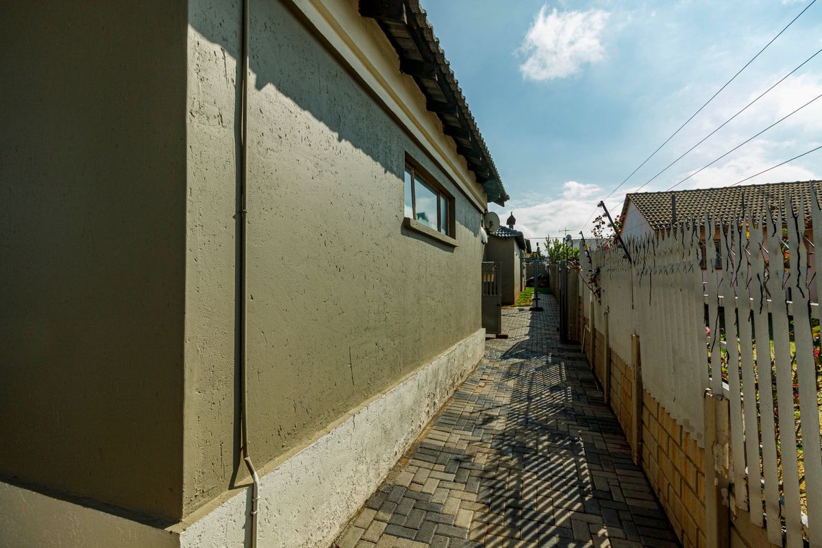 A side pathway featuring paved brickwork, a solid boundary wall with security spikes, and a view along the exterior of the property.