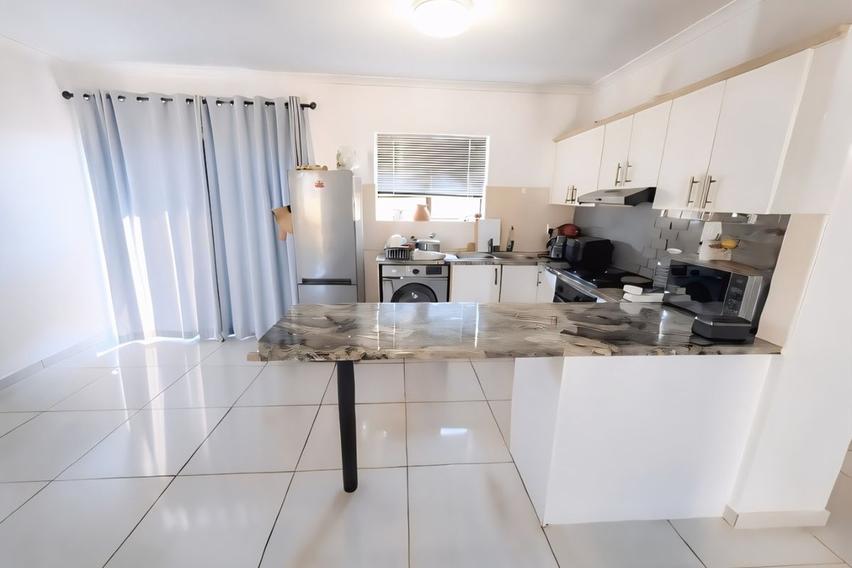 Open-plan kitchen and living area featuring white cabinetry, a marble-effect breakfast bar, tiled flooring, and direct access to a balcony through sliding doors.