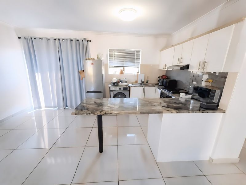 Open-plan kitchen and living area featuring white cabinetry, a marble-effect breakfast bar, tiled flooring, and direct access to a balcony through sliding doors.