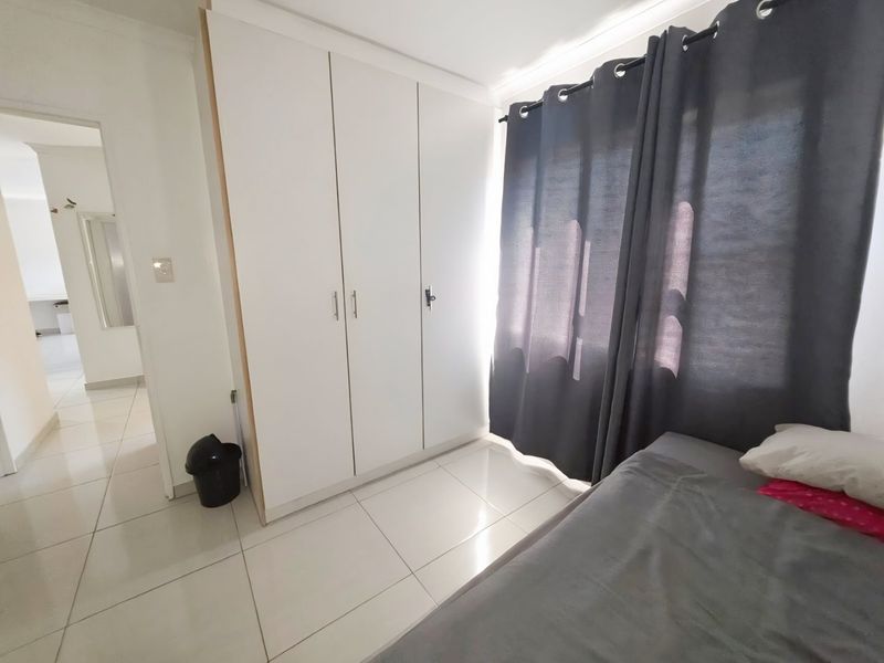 A bedroom featuring white built-in cupboards, neutral tiled flooring, and large windows with dark curtains, offering a clean and functional living space.