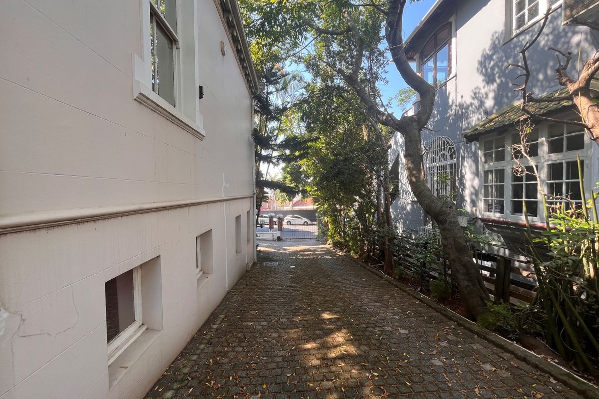 A narrow paved alleyway between two commercial building structures featuring mature trees and a secure gated entrance.