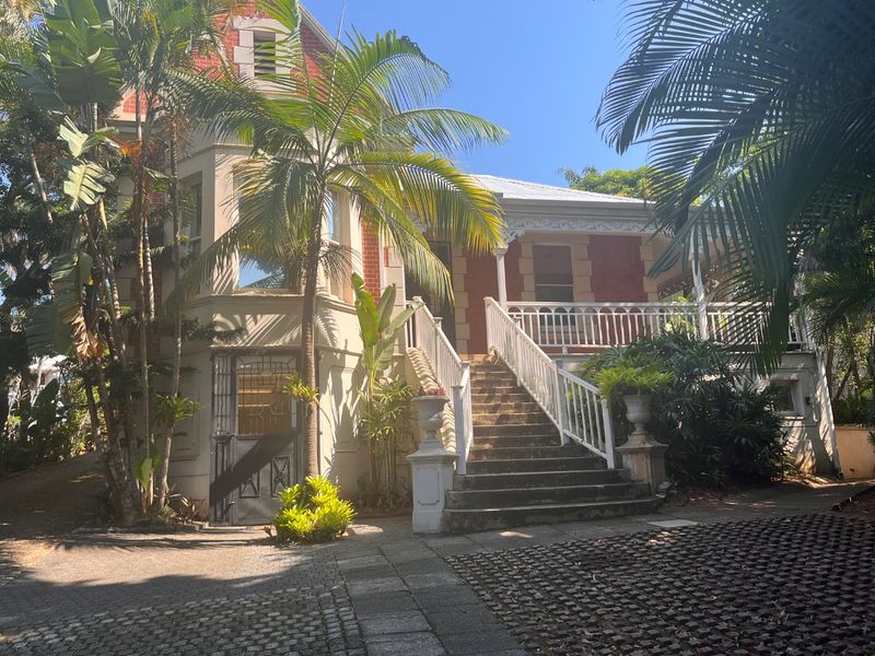 A charming historic commercial property featuring a classic brick facade, elevated veranda with balustrade, and a paved entrance area with mature landscaping.