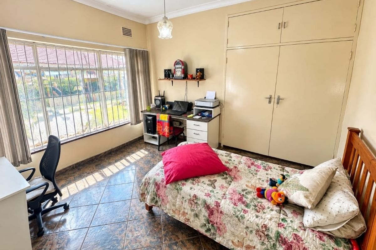 A bedroom featuring tiled flooring, built-in cupboards, and large windows with security bars, providing natural light and garden views.