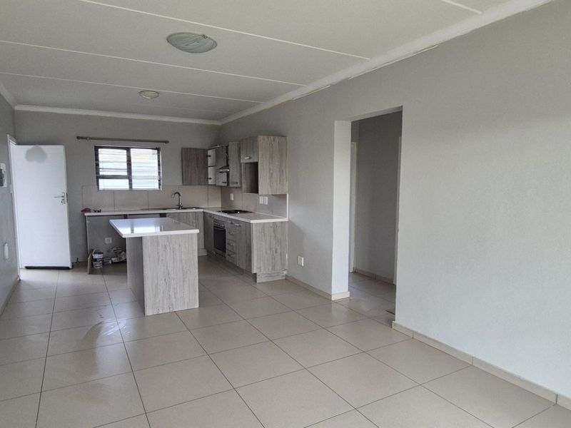 An open-plan living and kitchen area featuring modern wood-look cabinetry, a central island, built-in appliances, and neutral tiled flooring throughout.