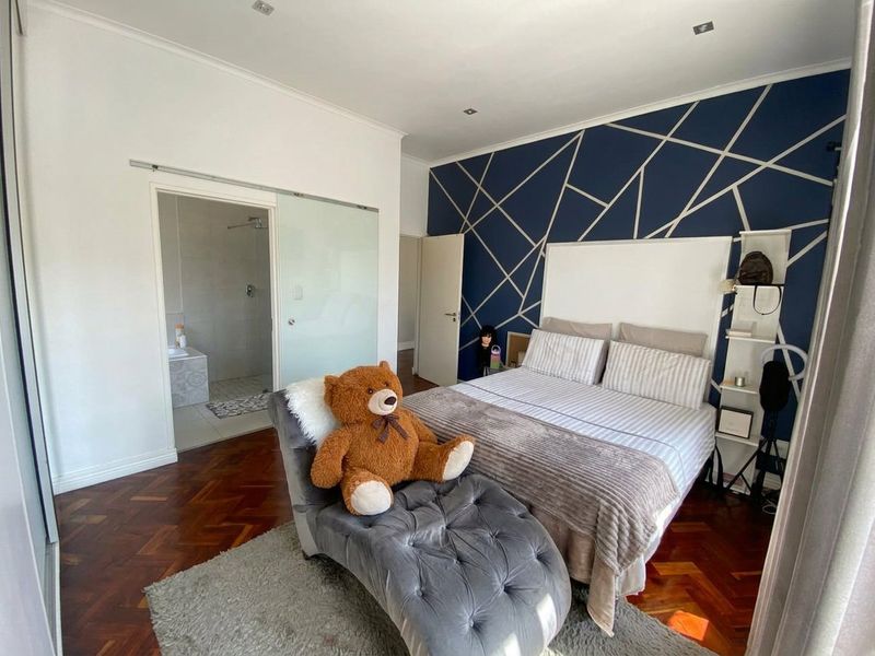Spacious bedroom featuring rich herringbone parquet flooring, a striking geometric accent wall, and direct access to an en-suite bathroom through a modern sliding glass door.