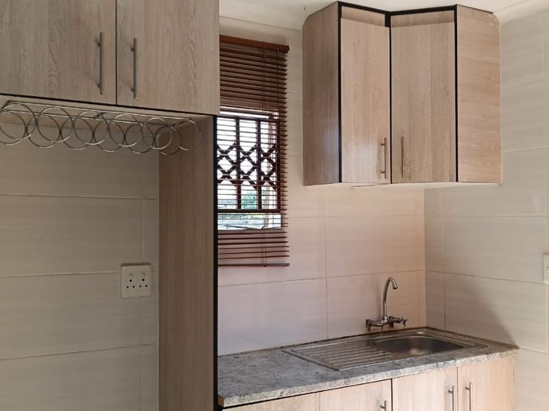 A compact kitchen featuring light wood-look cabinetry, a stainless steel sink, tiled walls, and a window with security features.