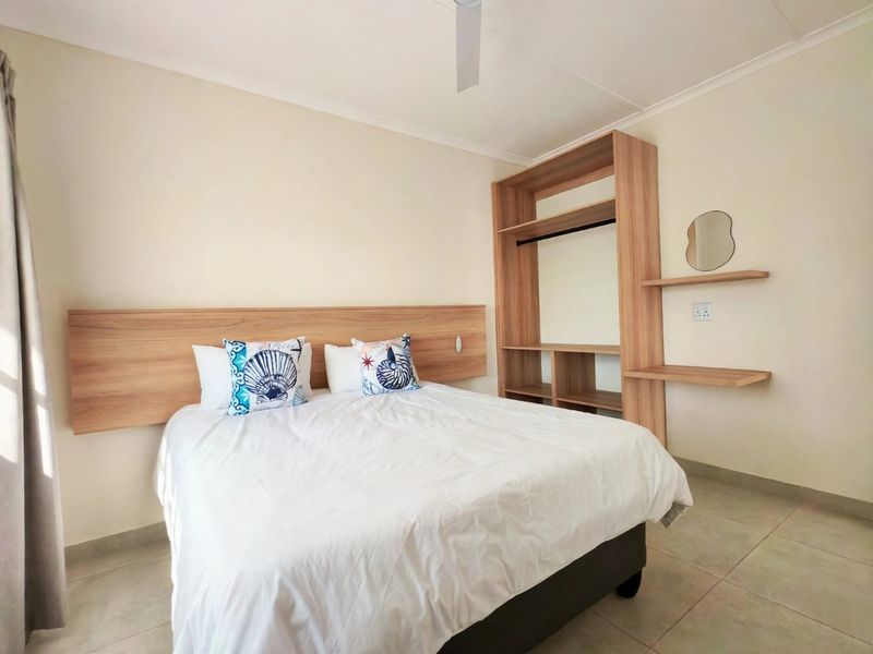 A well-maintained bedroom featuring a built-in headboard, integrated wardrobe with shelving, ceiling fan, and neutral tiled flooring.