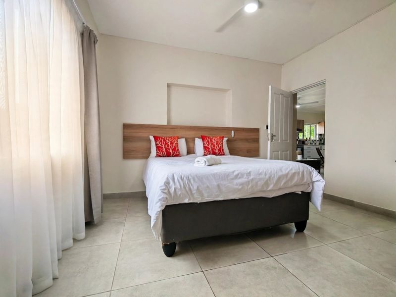 A well-maintained bedroom featuring neutral tiled flooring, a built-in wooden headboard, a ceiling fan, and a recessed wall niche, with an adjoining room visible.