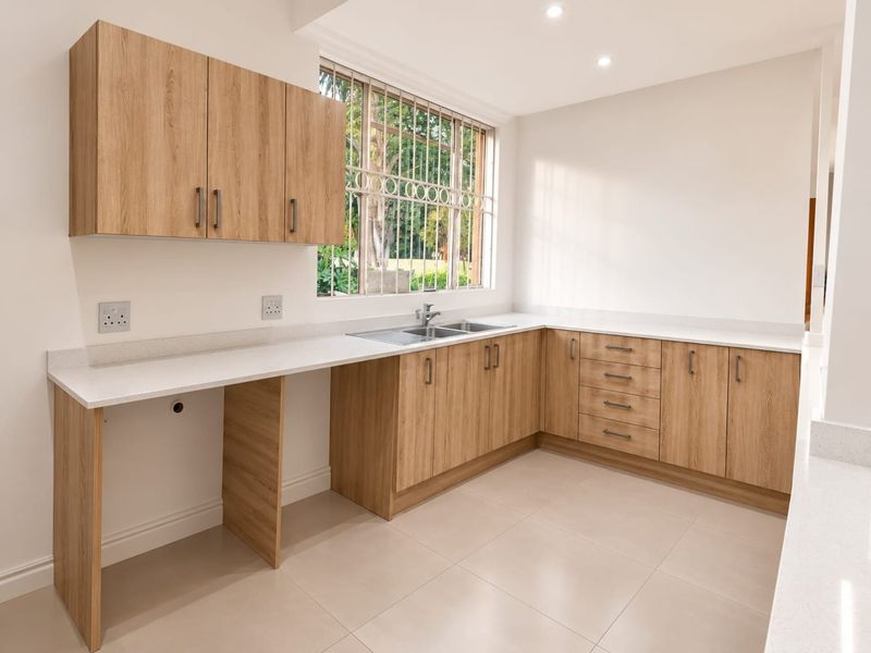 A clean, modern kitchen featuring light wood-finish cabinetry, stone countertops, a stainless steel sink, and neutral tiled flooring in an open, well-lit space.