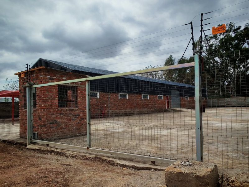 Industrial property featuring a secure gated entrance, brick guard house, and a large concrete yard enclosed by perimeter mesh fencing and electric security wires.