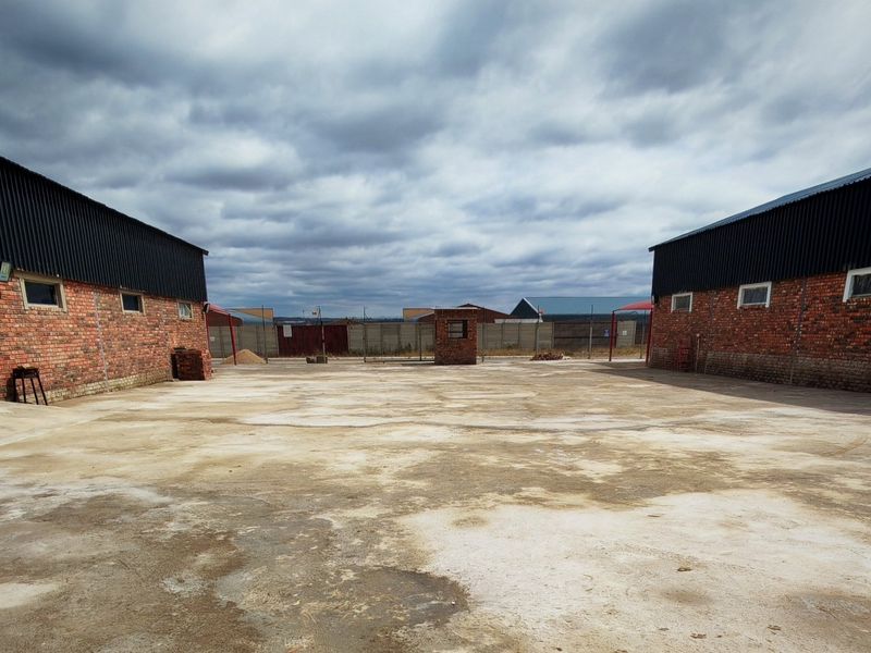 Spacious industrial yard area situated between two brick-built warehouse structures with corrugated metal roofing and perimeter security fencing.