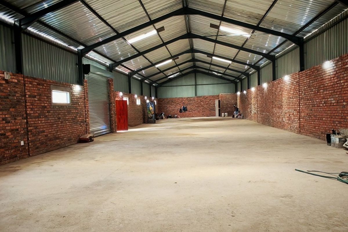 Spacious industrial warehouse unit featuring exposed brick walls, high-clearance steel truss roofing, concrete flooring, and a large roller shutter door.