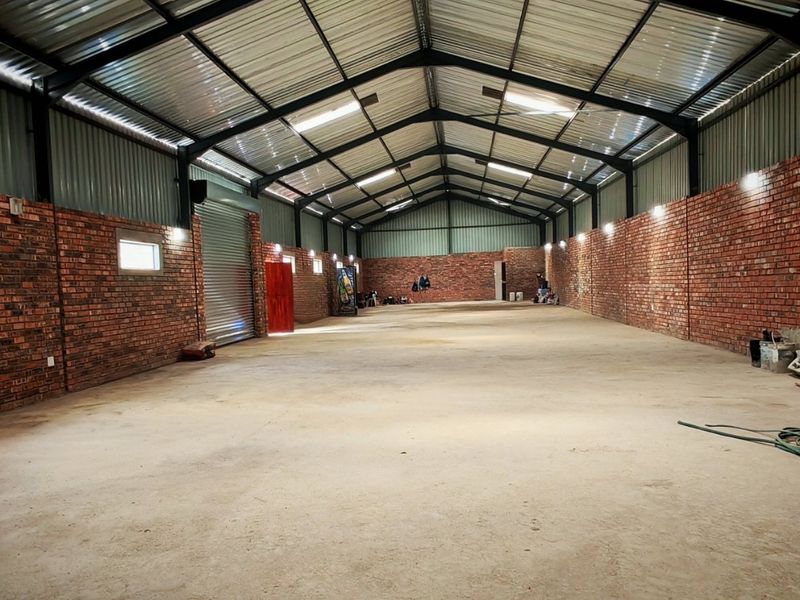 Spacious industrial warehouse unit featuring exposed brick walls, high-clearance steel truss roofing, concrete flooring, and a large roller shutter door.