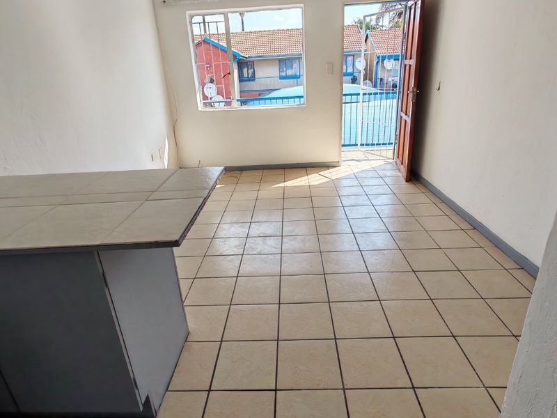 An empty open-plan living area featuring tiled flooring, a built-in breakfast counter, and direct access to an outdoor space through a security gate.