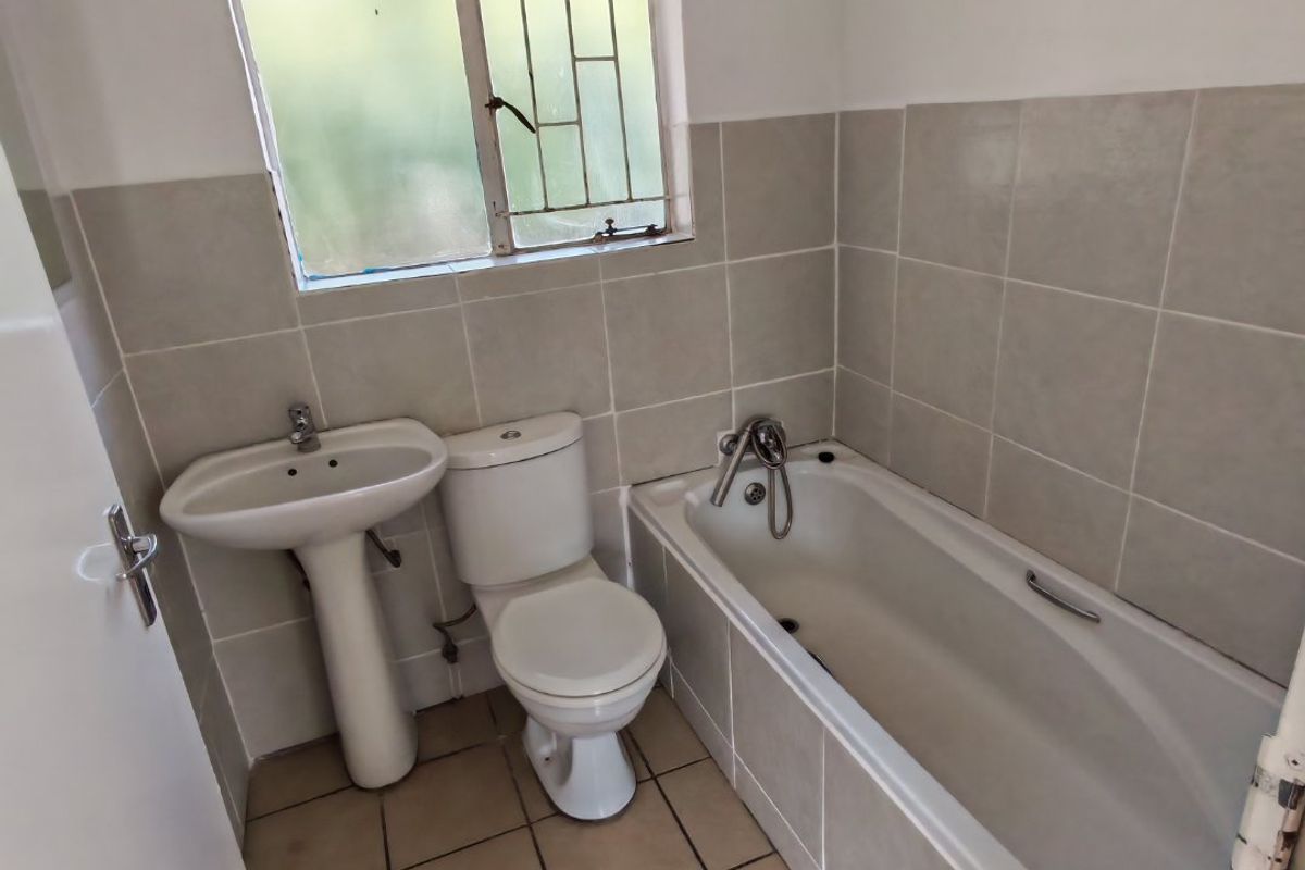A functional bathroom featuring a pedestal basin, toilet, and bathtub with tiled finishes and a window with security bars.