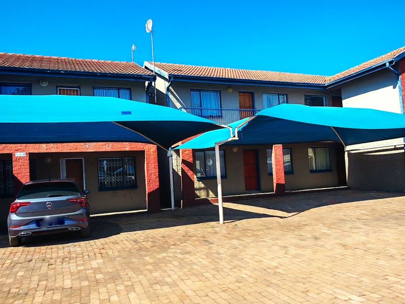Exterior view of an apartment complex featuring a paved driveway, covered parking bays with shade cloth, and brick facade finishes.