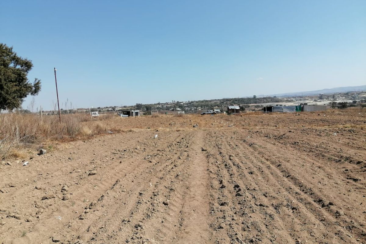 A wide-angle view of a vacant residential plot featuring cleared earth, perimeter fencing, and distant neighborhood structures under a clear sky.